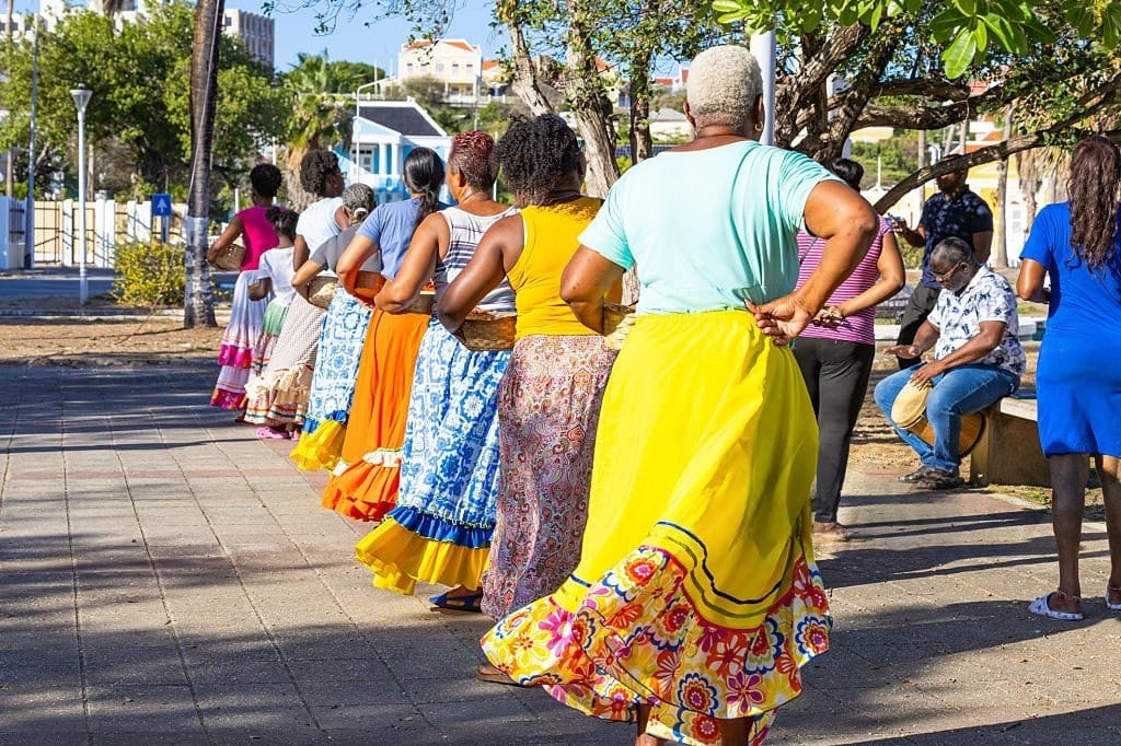 Femmes en jupes colorées dansant en file avec des paniers, un percussionniste assis à droite, à Willemstad