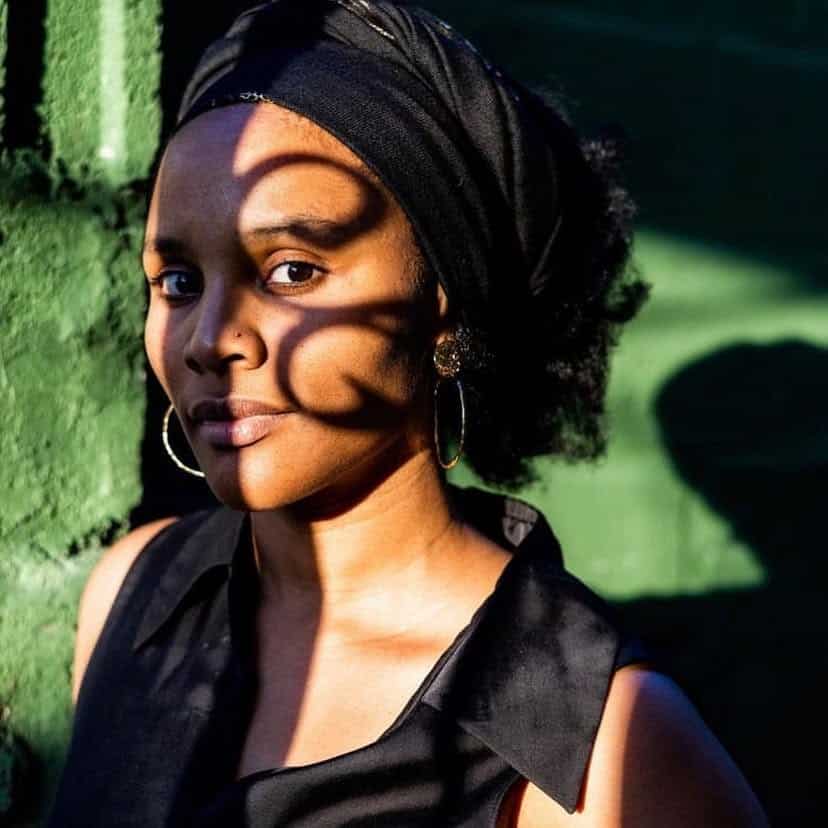 Portrait of a woman in a black headscarf, her face crossed by shadows, in front of a green wall