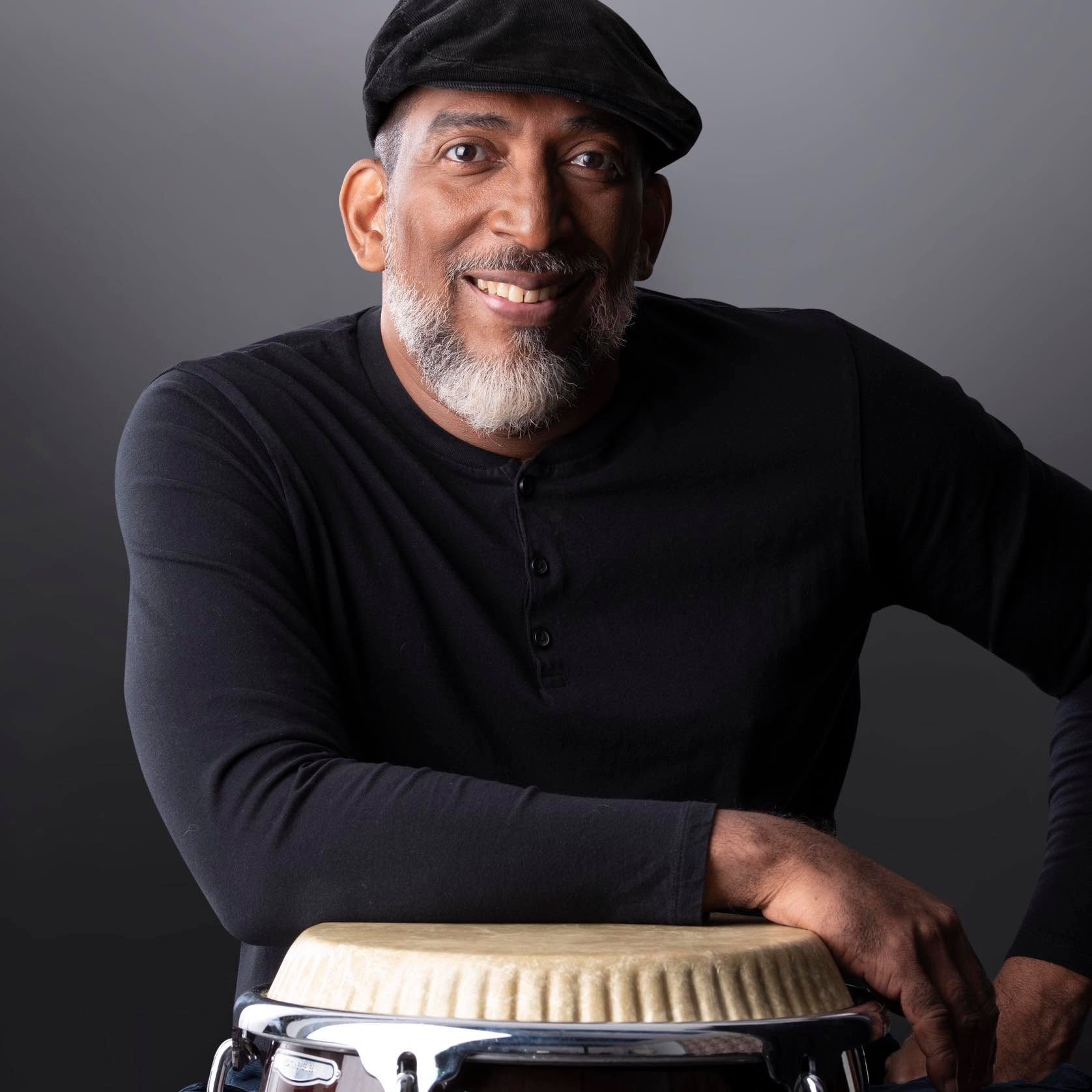 Chichí Peralta smiling, leaning on a conga, black cap, gray background