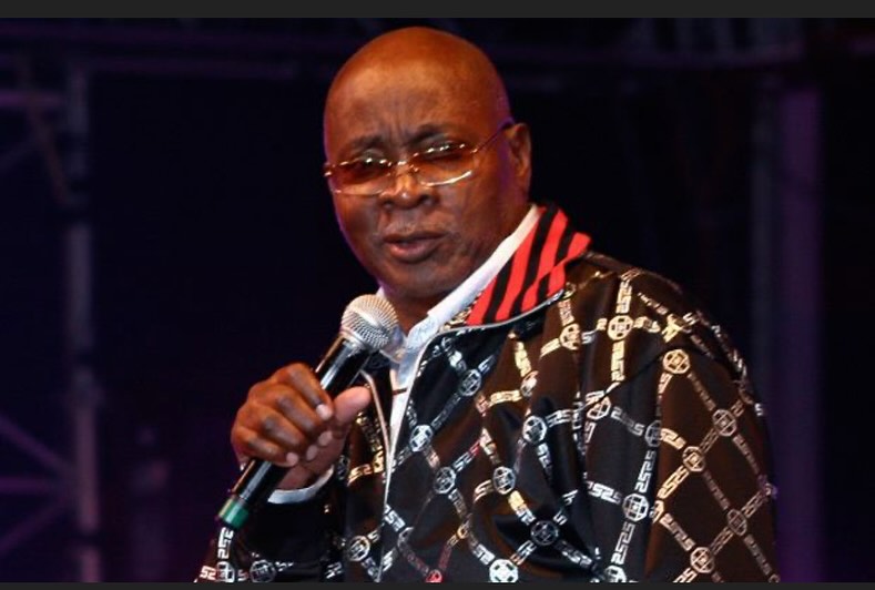Mighty Sparrow on stage, microphone in hand, wearing a black jacket with silver patterns