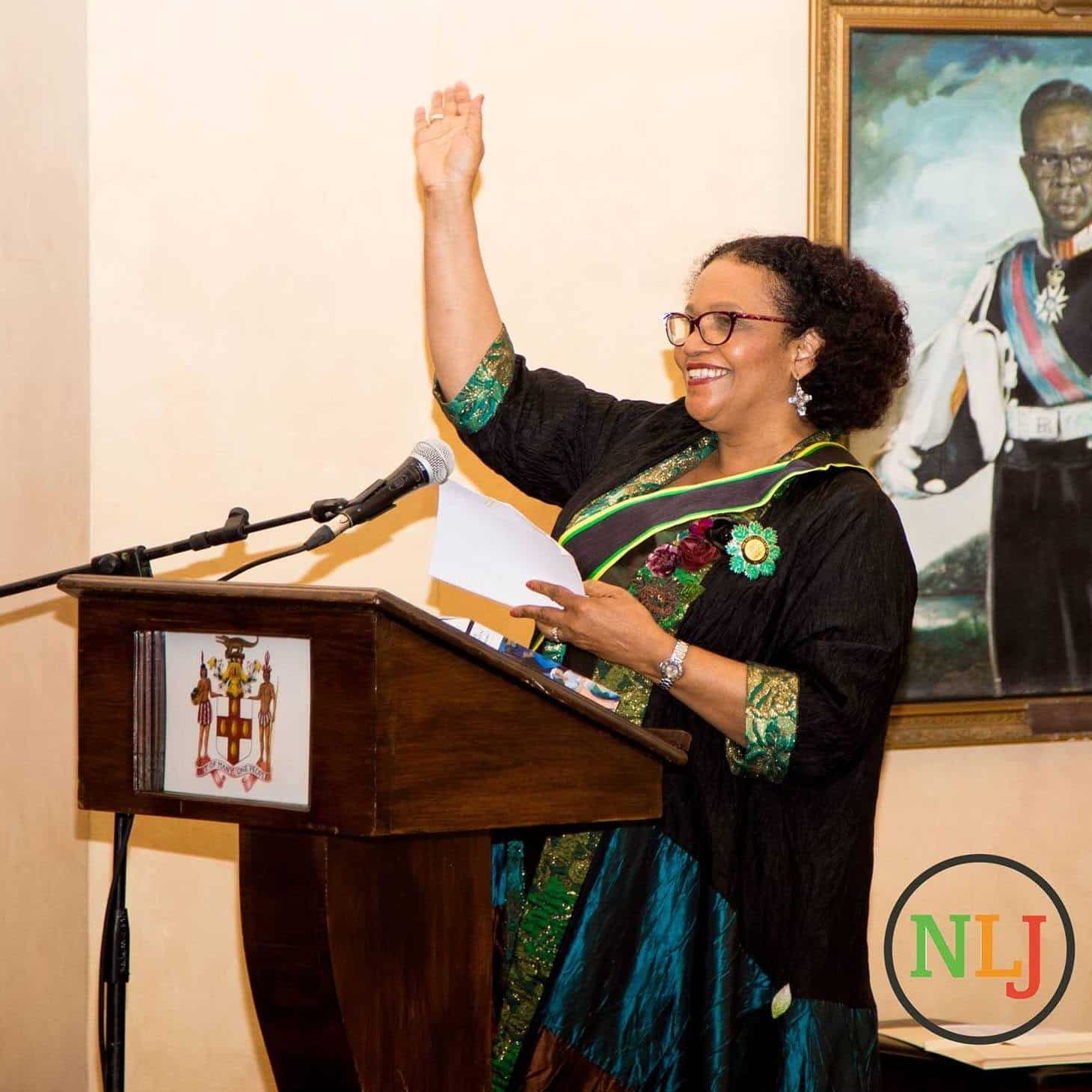 Lorna Goodison raising her hand at a lectern during a ceremony in Jamaica